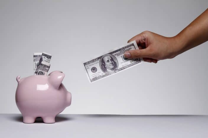 pink piggy bank with cash and hand with dollar bill reaching toward piggy bank