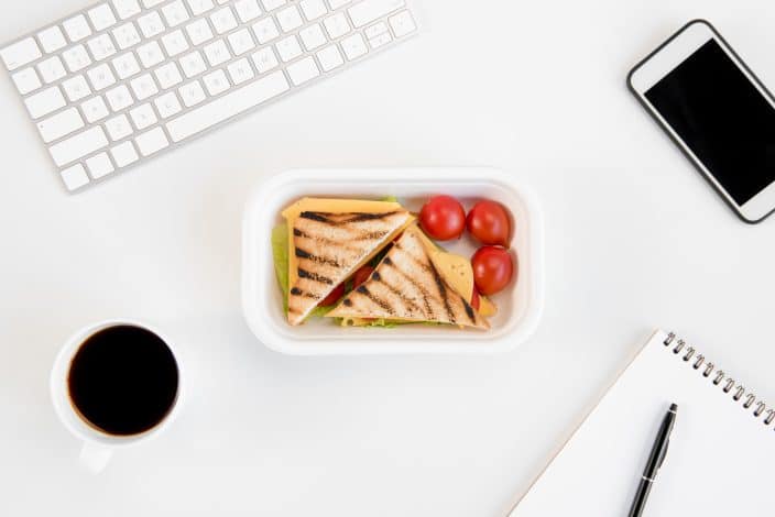 Top view of sandwiches with tomatoes in lunch box, notebook with pen, cup of coffee, smartphone and keyboard at workplace in order to cut eating out expenses