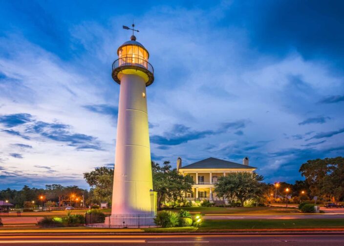 Picture of a lighthouse which has light glowing at the top during the evening, with trees and a big house in the background in Biloxi, Mississippi. 