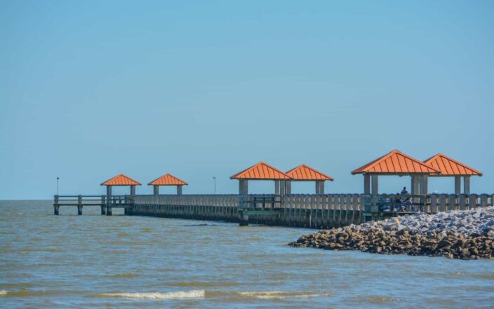 View of the shore with rocks, beachfront, blue skies and waves, with a dock with pergolas with orange roofs in the daylight in Gulfport,  Mississippi. 