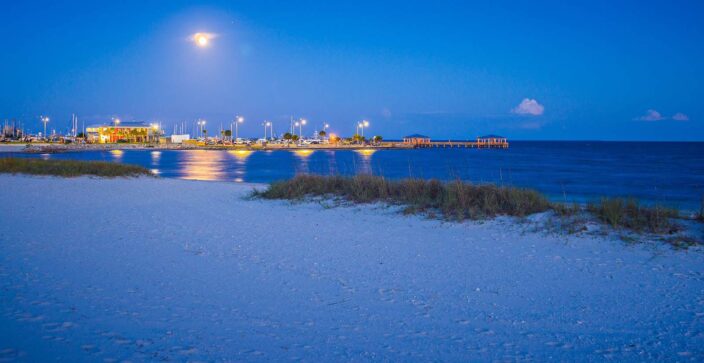 View of the beach with bluish sand, blue waters, and glowing lights of the pier and buildings in the background at night. 