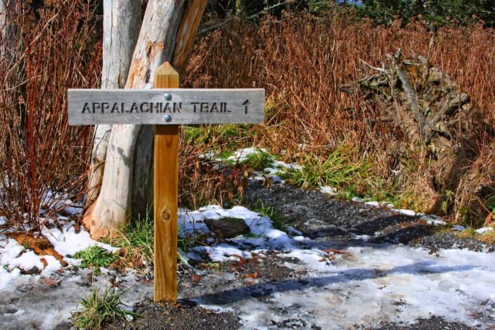 Signboard of the Appalachian trail with snow on the ground and brown bushes with a little bit of green grass in the background. 