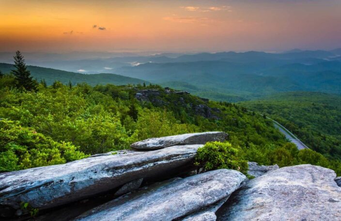 Image of the Blue Ridge Parkway in North Carolina with grey rocks, mountains, and lush green trees during the sunset. 
