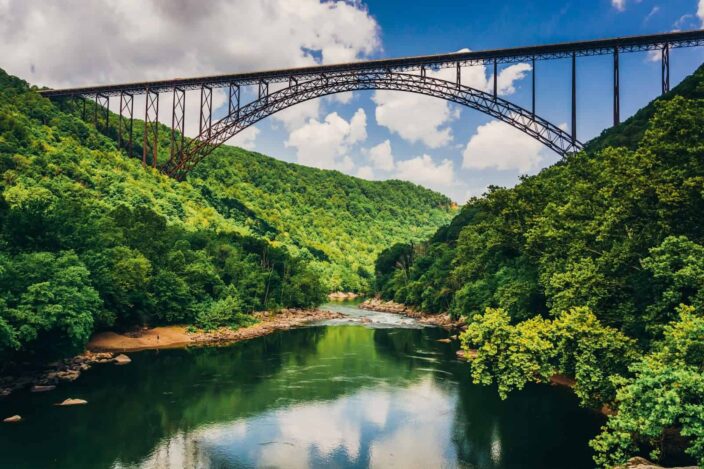 Picture of the River Gorge bridge with green hills on either side and river flowing underneath with blue skies and clouds. 