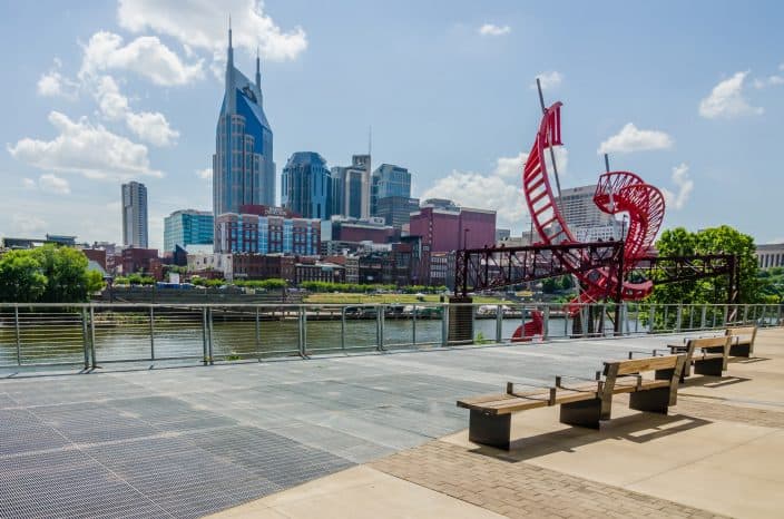 daylight view of Nashville city with skyscrapers, a red helix sculpture, benches, green trees, blue skies, and river in one frame. 