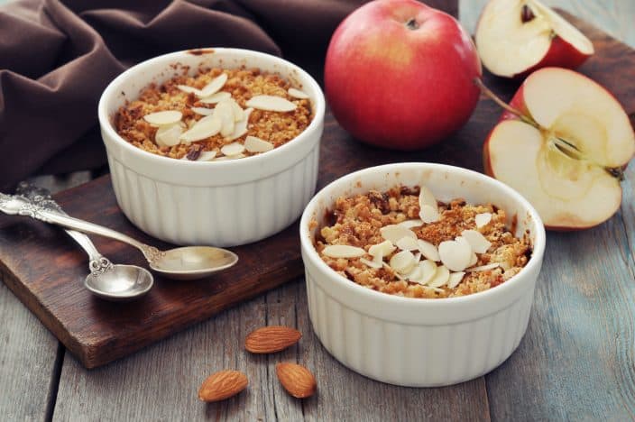 Apple crisp in  2 ceramic molds on a wooden cutting board on a wooden table with fresh apples, silver spoons, and almonds in the foreground.