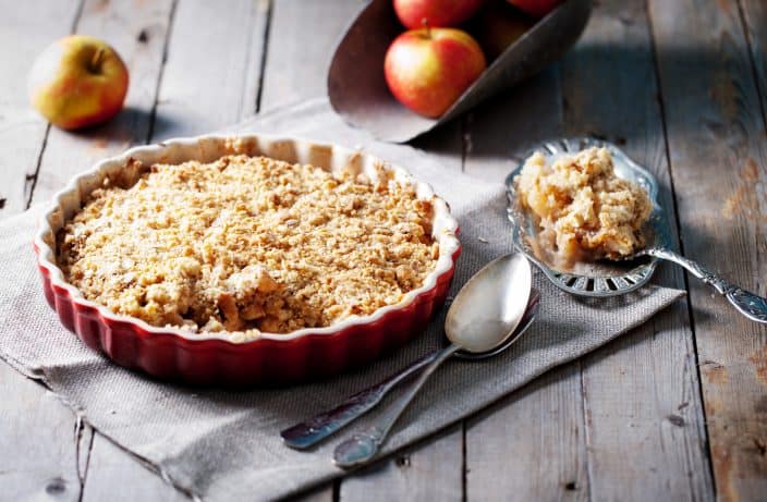 apple crisp in a red pie dish with silver spoons in front with a silver spoon filled with apple crisp on the side on a wooden background with whole apples in the background