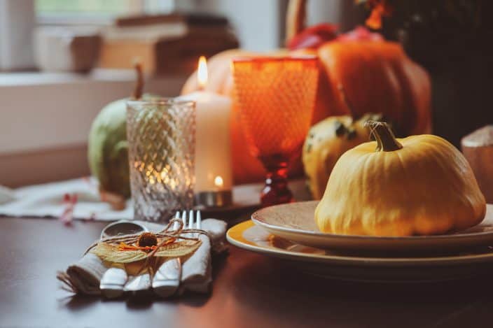 An autumn traditional table setting, with white and red candles, a squash sitting on a plate at a place setting and pumpkins on the table, an example of things to do in fall.
