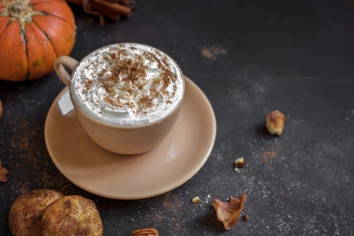A homemade Pumpkin Spice Latte topped with whipped cream and cinnamon with a pumpkin, snickerdoodle cookie, and spices next to it on a black background.