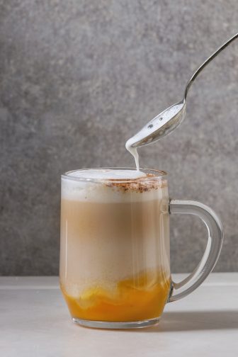 Glass of pumpkin layered spice latte with pumpkin puree, milk foam flowing from spoon and cinnamon standing on white marble table with grey wall at background