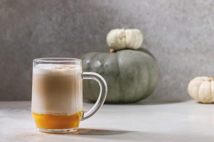 Glass of pumpkin layered spice latte with pumpkin puree, milk foam and cinnamon standing with decorative white pumpkins on white marble table with grey wall at background.