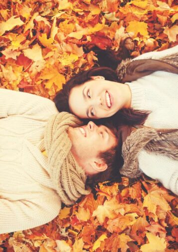 a couple laying in a pile of red, orange, and gold fall leaves on the ground in white sweater and smiling