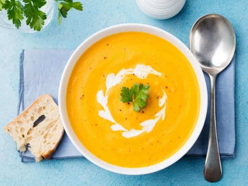 a bowl of pumpkin soup with sour cream on top and a blue background with a blue napkin, a silver spoon, a piece of bread, and a glass of water with parsley in it