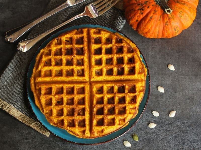 a pumpkin waffle on a teal plate, with a grey background and an orange pumpkin behind it with a silver fork and spoon and pumpkin seeds scattered around the plate, an example of a pumpkin, healthy recipes  