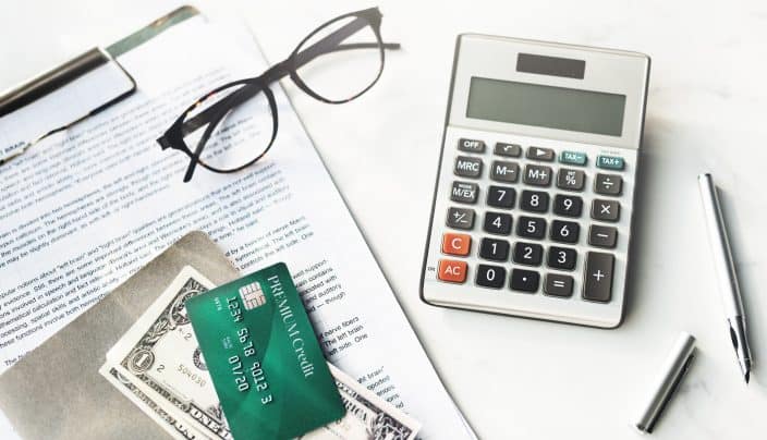 A calculator, papers, credit card, dollar bills, and glasses to be used for calculating and reducing debt