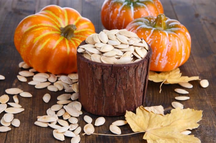 Pumpkin seeds in a wooden pot with orange pumpkins, pumpkin seeds, and maple leaves on the wooden table background