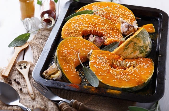 Cut open roasted pumpkin on a black baking sheet topped with white sesame seeds, garlic, and jars of orange and red spices in the background, an example of pumpkin healthy recipes