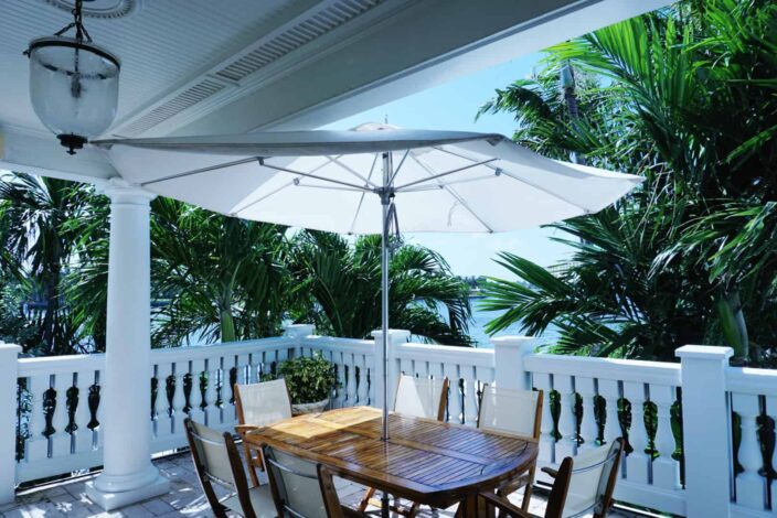 The Pillars, Fort Lauderdale balcony with stone railing, a white umbrella over a wooden table with white and wood woven chairs and a view of large green palm trees.