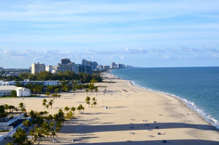 An aerial view of Fort Lauderdale beach with blue ocean, skies and beach with green palm trees and sky scrapers in the distance. 