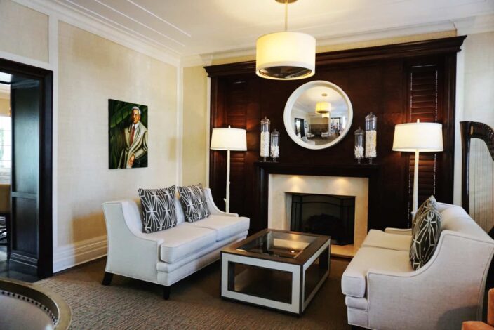 a view of the seating and lounge area of The Pillars, Fort Lauderdale with beige couches, a mahogany wall accent with a fireplace, colorful carpet, a painting on the wall, yellow-beige walls, a glass coffee table, and white standing lamps next to the couch. Behind the couch on the right is a harp. 