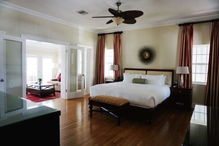 a view of a suite at The Pillars, Fort Lauderdale with mahogany wood floors, bed frame and side tables. The walls are a light yellow beige color, with double doors leading to the living area of the suite with a red rug and a beige couch, and wood table. 