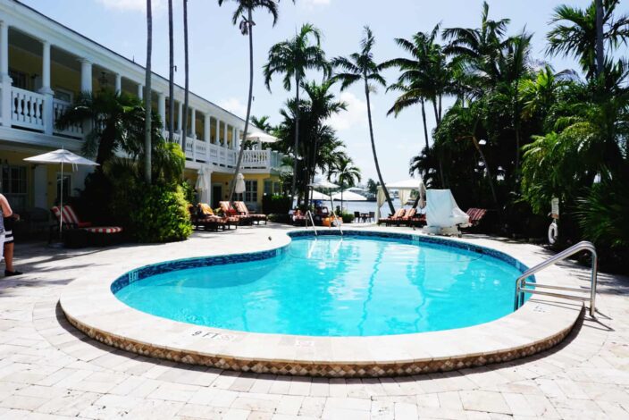 a view of the pool with cobble stone paving at The Pillars Hotel in Fort Lauderdale with many green palm trees, red and white striped lounge chairs, and a view of blue skies and the waterfront in the background 