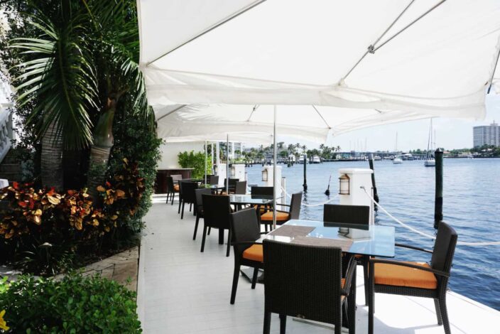 an image of the view at the Secret Garden restaurant at The Pillars hotel featuring dining chairs and tables along the Intercoastal Waterway with white umbrellas above and greenery and palms on the side