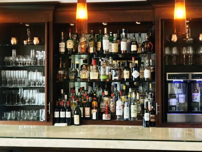 The Pillars, Fort Lauderdale bar with many bottles of alcohol lined up against a mirrored wooden cabinet with alcohol glasses on the left side and more glasses on the right with a wine cooler 