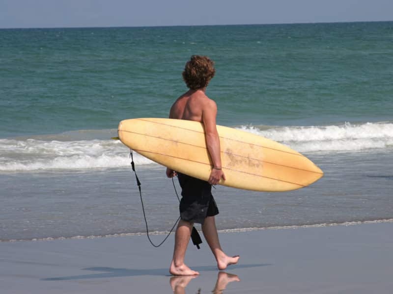 man on beach carrying a tan surfboard, walking on the damp sand with waves breaking on shore