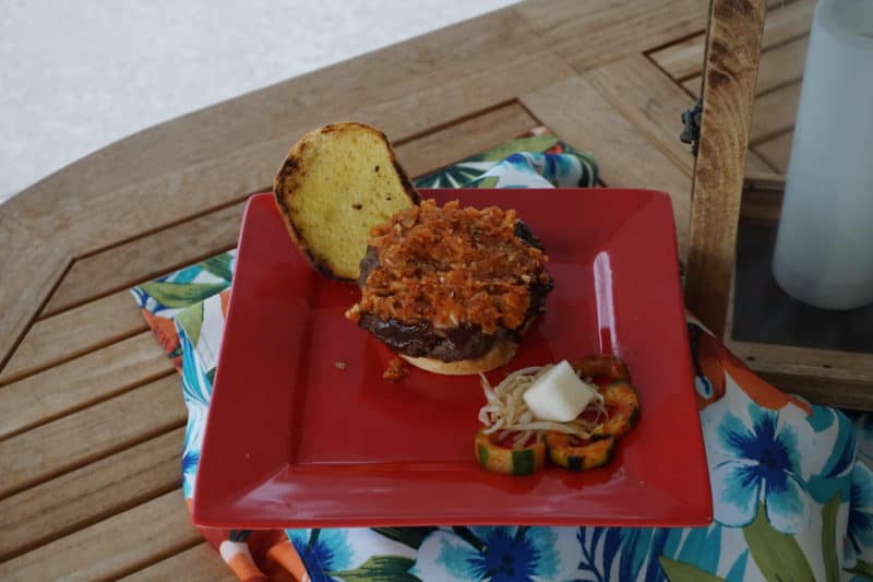 Red plate, toasted hamburger bun, hamburger with kimchi slaw, and cucumber and radish side dish