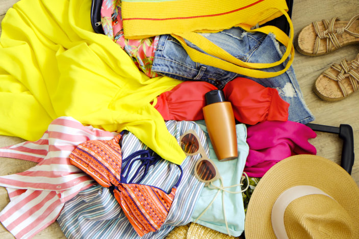 Open suitcase with pile of unfolded clothing on the floor. Woman packing for tropical vacation concept. Multiple unpacked female clothing items prepared for travel. Background, close up, copy space.