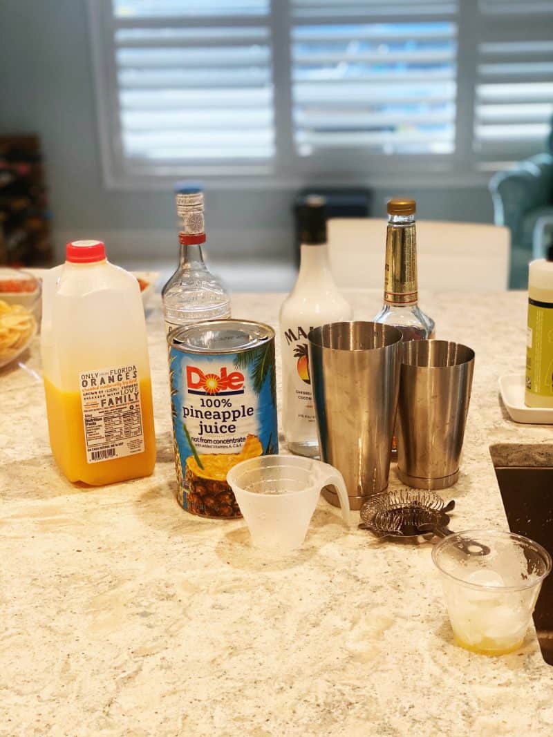 Ingredients to make a Goombay Smash sitting on a counter top including orange juice, pineapple juice, Malibu rum, a plastic cup with ice and orange juice in it, a measuring cup,  a strainer, and a cocktail shaker.