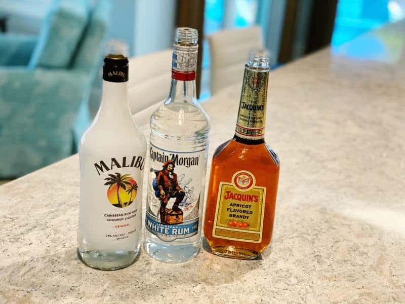 The ingredients to make a goombay smash cocktail including a bottle of Malibu rum, Capitan Morgan White Rum, and apricot brandy, sitting on a countertop