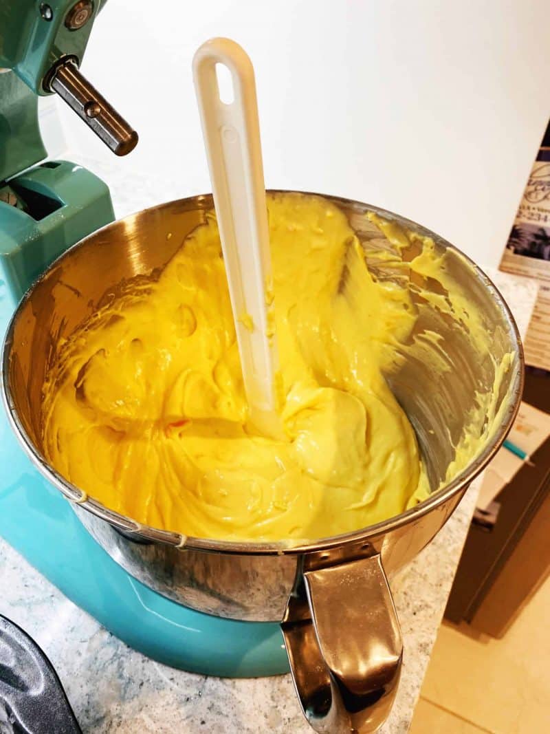 mixing yellow batter in an teal electric stand mixer on a beige countertop with a rubber spatula in the batter to make a Tortuga rum cake 