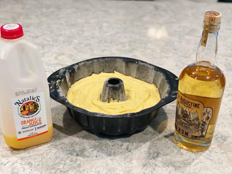 batter for rum cake in a Bundt cake pan with Natalie's Orange Juice on the left and a bottle of rum on the right sitting on a kitchen counter top