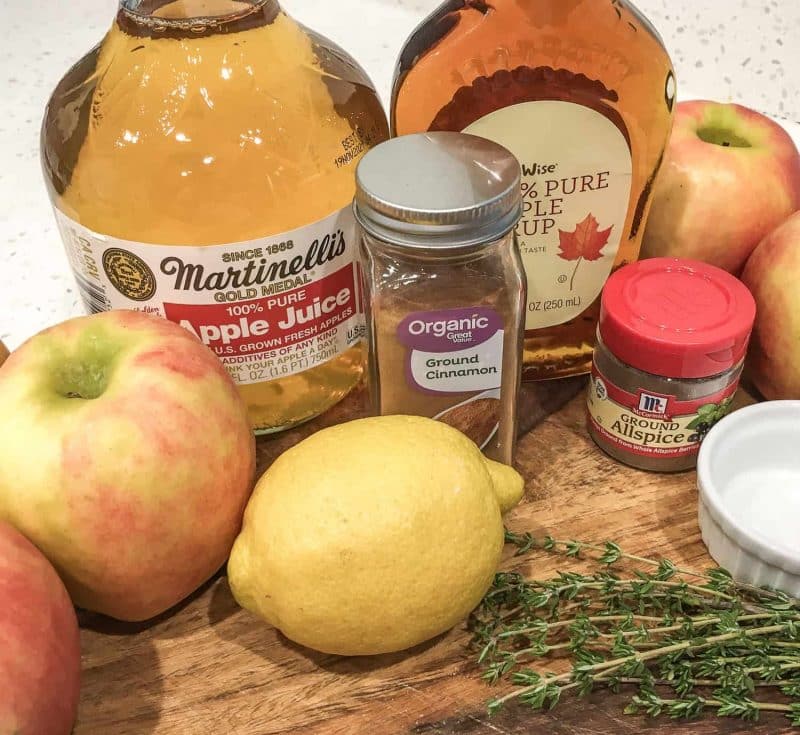 a jar of apple juice, cinnamon, allspice, apples, a lemon, and thyme on a wooden cutting board on a white counter