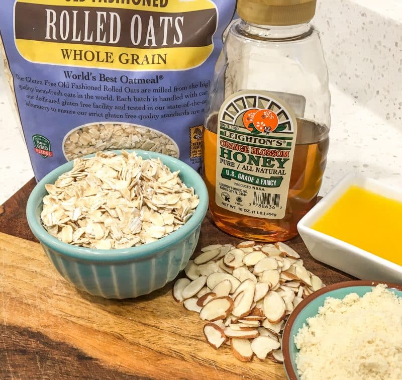 Topping ingredients for baked apple cinnamon crisp featuring a bag of rolled oats, honey, almonds in a teal ramekin, melted butter in a white ramekin, and almond flour in a teal plate on a wooden cutting board on the counter 