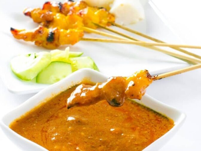 sauce in a white dish with spicy chicken satay skewers on a white plate with sliced cucumber