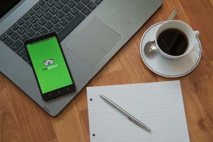 trip advisor app open on a cell phone with a laptop, coffee, and a notepad for taking notes on places to stay during your vacation