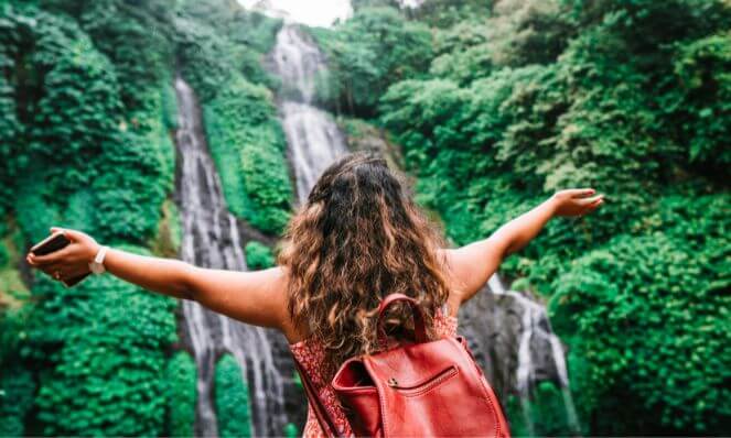women with open arms looking at waterfall while traveling with a well packed red suitcase