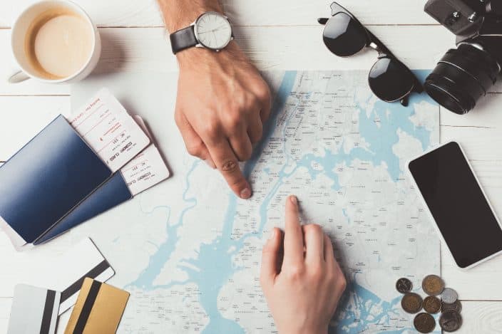 a man's and woman's on a map with passports, sunglasses, a camera, phone, money, and credit cards planning out a trip