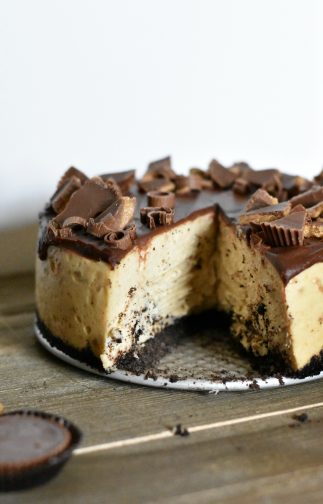Peanut butter cheesecake with a slice taken out, topped with chocolate ganache and Reese's pieces.