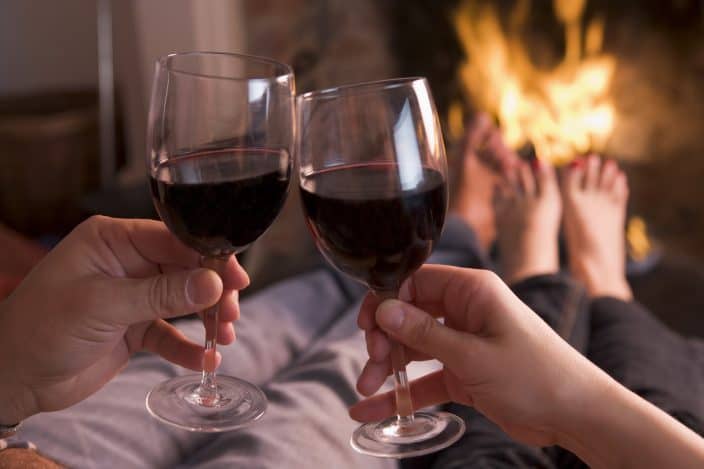 two people laying down in from of a fire place drinking two glasses of red wine. 