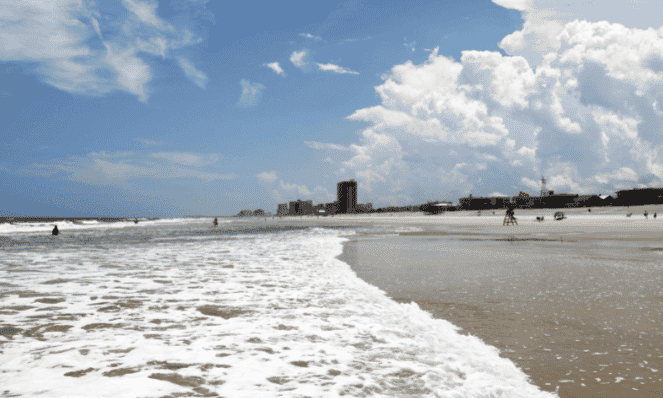 dog friendly Jacksonville beach with white sands and water lapping on the beach, sky rises viewed in the distance, and people enjoying their day playing on the beach with the blue skies and white clouds
