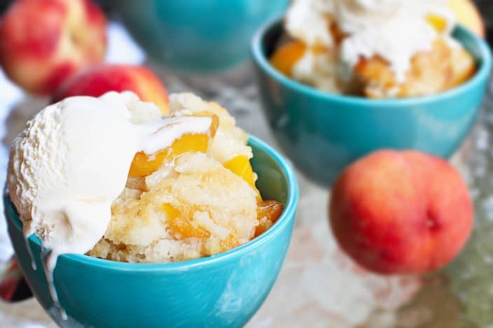 Fresh peach cobbler served in a teal bowl with vanilla ice cream on top.