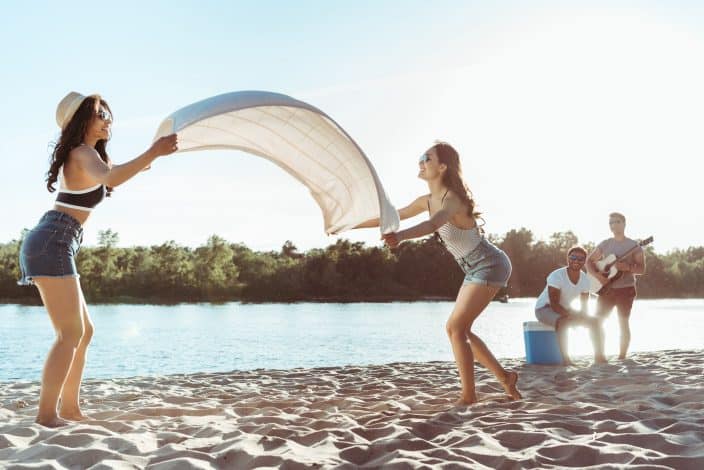 two girls laying down the blanket on the sand for their beach day with the river and two boys sitting on a cooler in the background looking at them. 