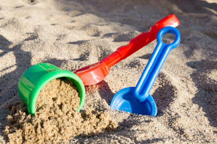 A picture of sand toys, a green basket, and two shovels red and blue in the sand. 