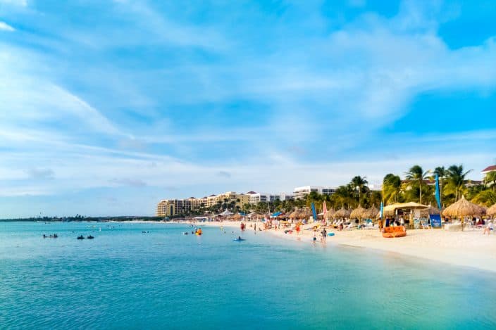 Palm beach in Aruba with blue waters, high rise hotels, and people on the beach swimming and sunbathing.