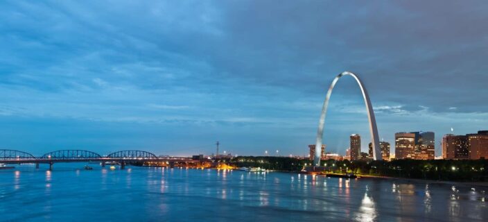 view of the Bay St. Louis, Mississippi in night time with bright lights and the glowing skyline, with the bay and a bridge in the background. The arc sculpture is also in the background. 