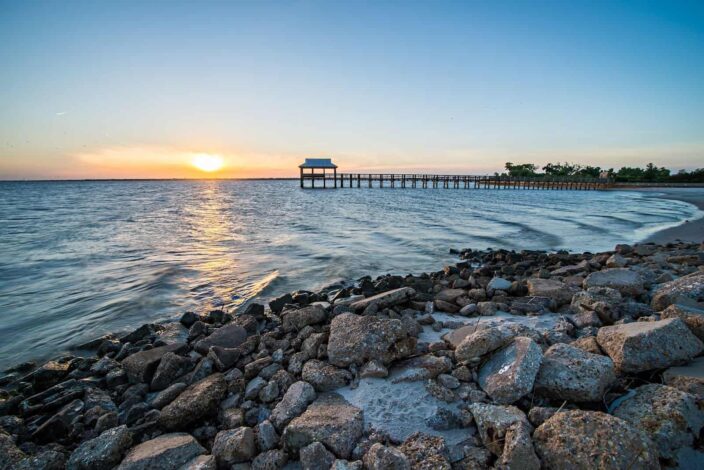  View of the shore at Pass Christian with rocks, beachfront, wood pier and pergola, and the sunset all in one frame.